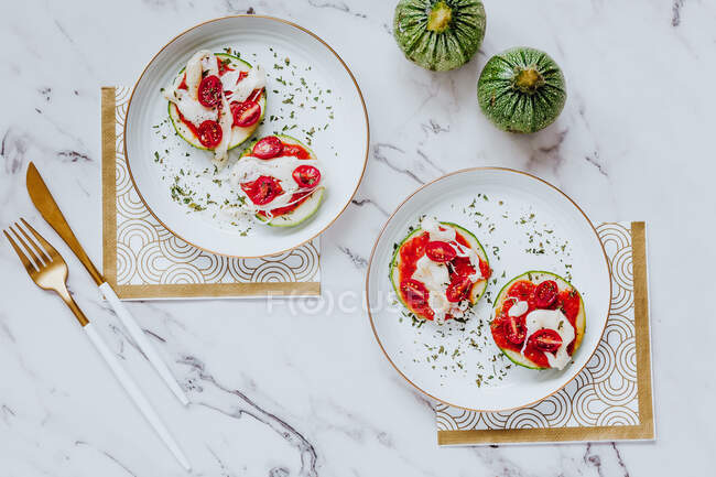 Сверху вкусные здоровые бутерброды с ломтиками свежих зеленых цуккини с расплавленной моцареллой и помидорами черри на белых тарелках с золотыми столовыми приборами — стоковое фото