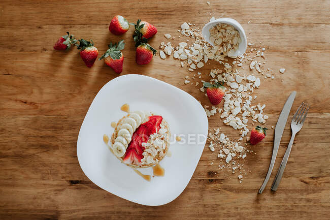 Pilha de panquecas saborosas servidas em prato com pedaços de banana e morango e mirtilos frescos com flocos de coco durante o café da manhã — Fotografia de Stock