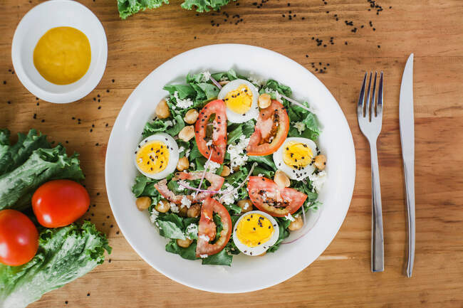 Top view of bowl of tomato and lettuce salad with chickpeas and boiled eggs placed on table with silverware and sauce — Stock Photo
