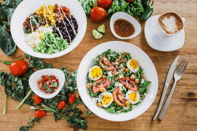 Taças e pratos de deliciosos pratos saudáveis com legumes e carne colocados perto de xícaras de café e molhos com tomates e ervas na mesa — Fotografia de Stock
