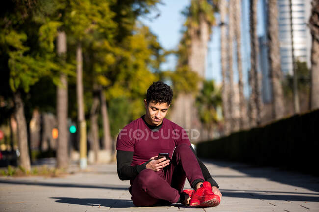 Atleta hispano sin camisa feliz sentado cerca del tronco del árbol y el teléfono inteligente de navegación mientras descansa durante el descanso en el entrenamiento de fitness en el parque - foto de stock