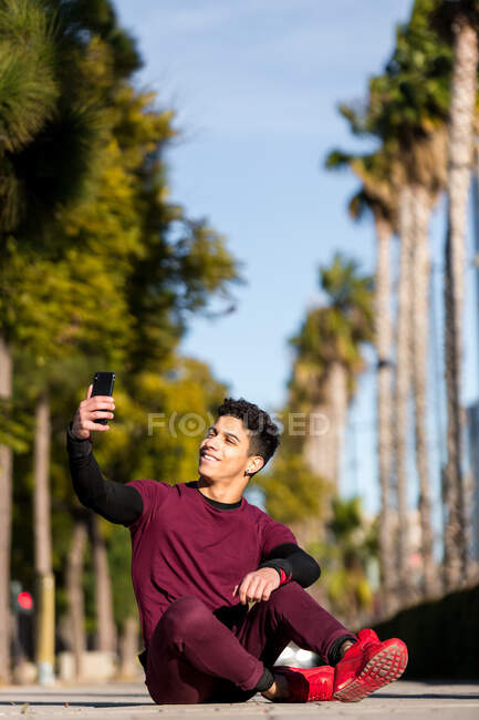 Веселый молодой этнический парень в спортивной одежде улыбается и делает селфи, сидя на пути во время тренировки в солнечный день в городе — стоковое фото