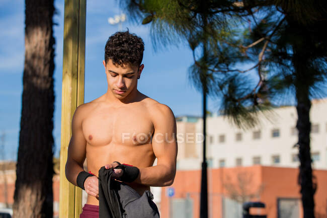 Shirtless ragazzo ispanico esaminando t shirt mentre in piedi sulla strada della città durante l'allenamento di fitness nella giornata di sole — Foto stock