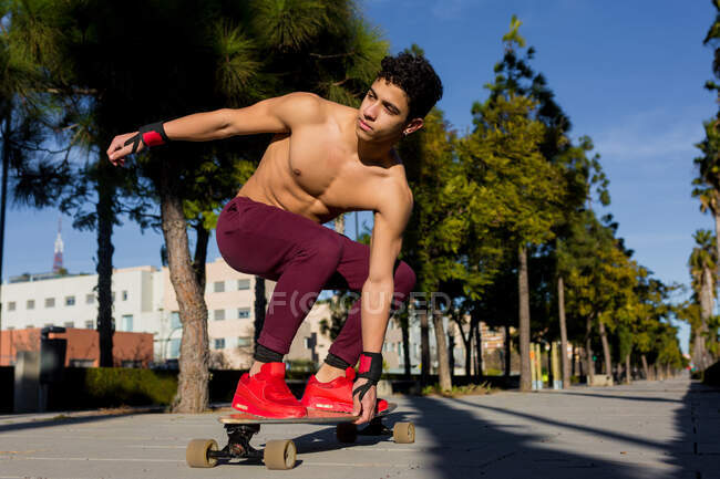 Full body young Hispanic man with backpack riding skateboard on sidewalk on sunny day on city street — Stock Photo