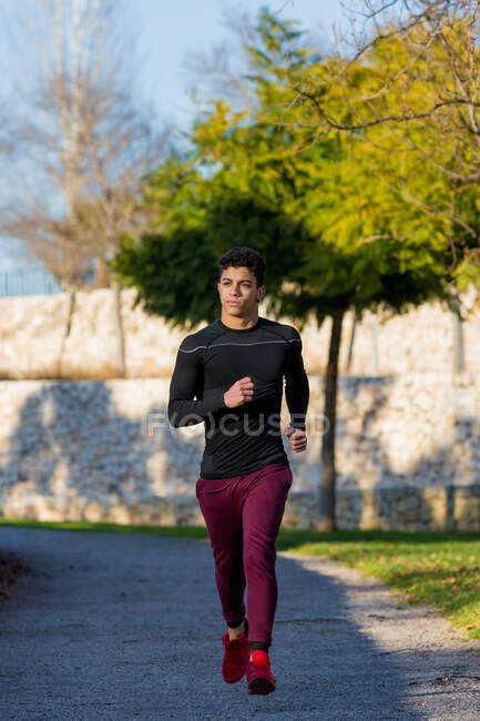 Atleta masculino étnico de comprimento total correndo no caminho durante o treino de fitness no dia ensolarado no parque — Fotografia de Stock