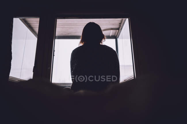 From bellow silhouette of anonymous lonely woman sitting on bed against window in dark room at home — Stock Photo