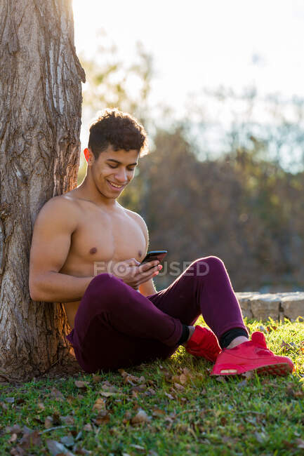 Feliz atleta hispânico sem camisa sentado perto do tronco da árvore e smartphone de navegação enquanto descansa durante a pausa no treinamento de fitness no parque — Fotografia de Stock
