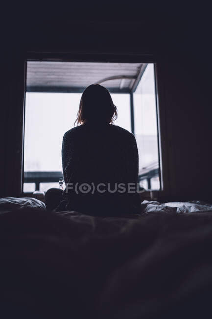 De silhueta abaixo de mulher solitária anônima sentada na cama contra a janela no quarto escuro em casa — Fotografia de Stock