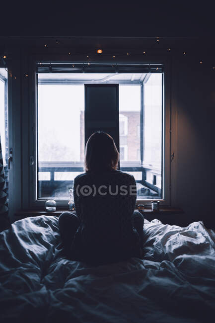 Vista posteriore della giovane donna depressa seduta sul letto in camera da letto scura a guardare lo specchio a casa — Foto stock