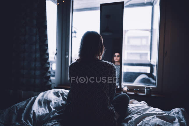 Back view of depressed young female sitting on bed in dark bedroom at looking at mirror at home — Stock Photo