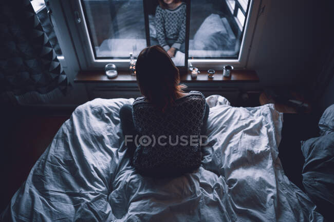 Back view of unrecognizable depressed young female sitting on bed in dark bedroom looking at mirror at home — Stock Photo