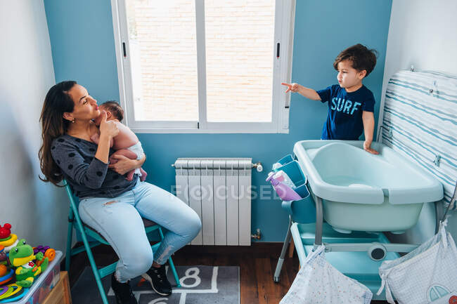 Alegre joven madre con bebé sentado en la silla y hablando con su hijo pequeño preparándose para bañar al recién nacido - foto de stock
