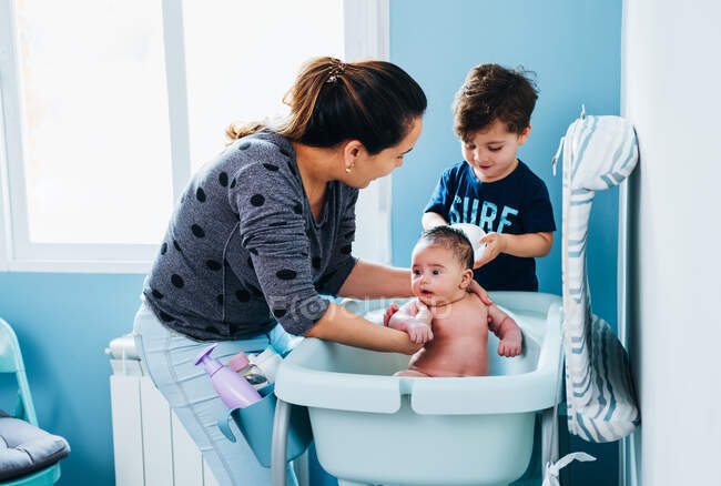 Adulto mulher carinhosa em suavemente lavar o bebê no banho de bebê no banheiro acolhedor, enquanto o pequeno filho ajudando a mãe e segurando tigela de água morna nas mãos — Fotografia de Stock