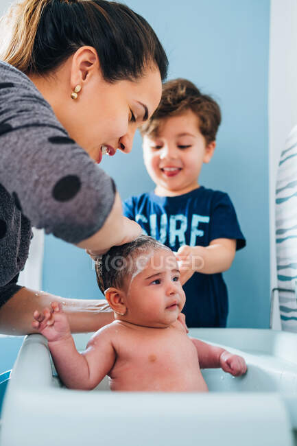 Доросла доглядаюча жінка в ніжному митті дитини в дитячій ванні в затишній ванній кімнаті, а маленький син допомагає мамі і тримає миску теплої води в руках — стокове фото