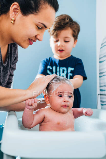 Adulto mulher carinhosa em lavar suavemente o bebê no banho do bebê no banheiro acolhedor, enquanto o pequeno filho ajudando a mãe — Fotografia de Stock