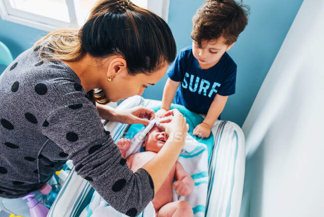 Desde arriba de la mujer cuidadosa y el pequeño hijo en ropa casual envolver bebé lindo llorando después del baño en el vivero de luz azul - foto de stock