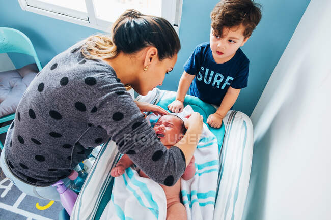 From above of careful female and little  son in casual wear swaddling cute crying baby after bath in blue light nursery — Stock Photo