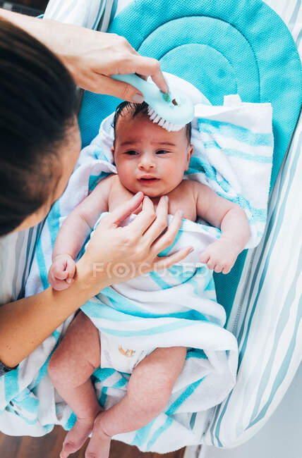 Женщина на поле нежно расчесывает симпатичного новорожденного, завернутого в голубое одеяло маленькой белой расчёской за ванной, в то время как малыш внимательно смотрит на маму и лежит на столе для переодевания. — стоковое фото