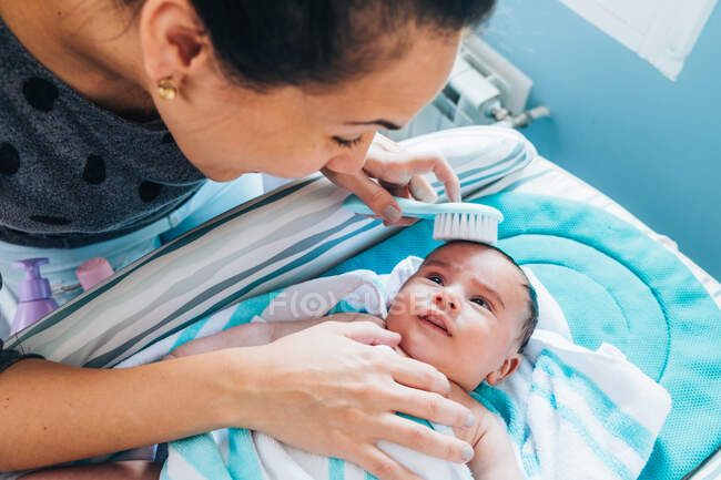 Mulher de colheita penteando suavemente bonito recém-nascido envolto em cobertor azul por pequena escova de cabelo branca após o banho, enquanto o bebê cuidadosamente olhando para a mãe e deitado na mesa de mudança — Fotografia de Stock