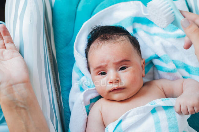 From above cute baby in blue bath towel lying on changing table and looking at camera while crop mother holding comb in hand — Stock Photo
