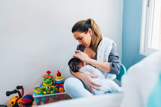 Mother breastfeeding infant while sitting on chair with legs crossed and patting baby on back in cozy nursery — Stock Photo