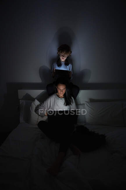 Focused boy and adult woman in sleepwear sitting cross legged on bed and using modern gadgets while relaxing in dark bedroom at home together — Stock Photo