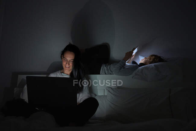 Доросла жінка і хлопчик в піжамі відпочивають на ліжку і переглядають цифрові пристрої в темній спальні вдома — стокове фото