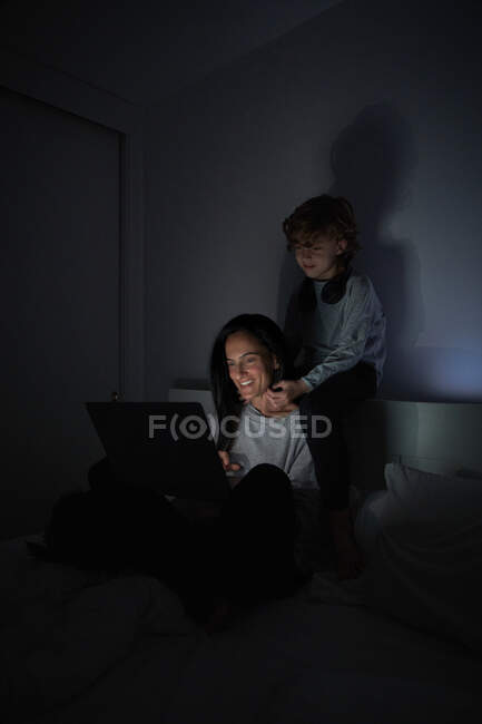 Niño abrazando alegre mujer adulta sonriendo y navegando portátil en habitación oscura en casa por la noche - foto de stock