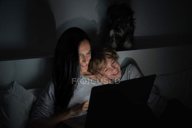 Happy boy leaning on shoulder of adult woman while lying on bed and watching movie on laptop at night at home together — Stock Photo