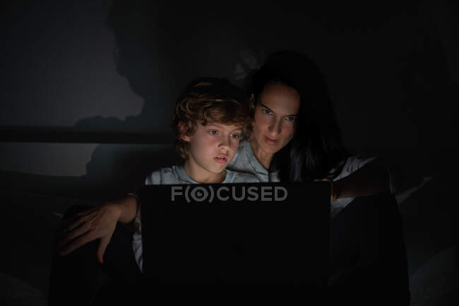 Boy leaning on shoulder of adult woman while lying on bed and watching movie on laptop at night at home together — Stock Photo