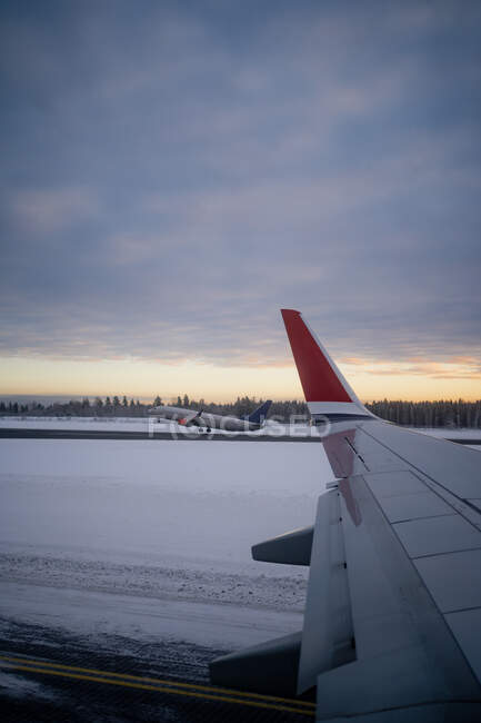 Flügel eines modernen Flugzeugs auf schneebedecktem Gelände gegen dunklen Wald am Horizont und Flugzeuge, die von der Landebahn in den grauen Himmel starten, bei bewölktem Wetter in der Abenddämmerung in Norwegen — Stockfoto