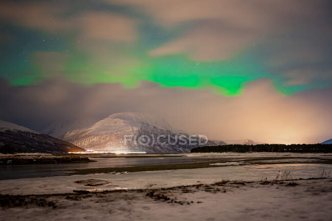 Pintoresco paisaje con asentamiento iluminado en la orilla del estrecho a pie de montañas nevadas bajo el cielo estrellado nublado con increíbles auroras boreales verdes en Lofoten - foto de stock