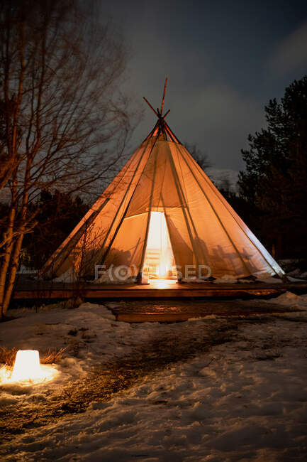 Chemin de randonnée enneigé menant à une tente de tipi en forme de cône confortable avec feu à l'intérieur sur un endroit en bois parmi les arbres persistants et sans feuilles dans les bois en soirée en Norvège — Photo de stock