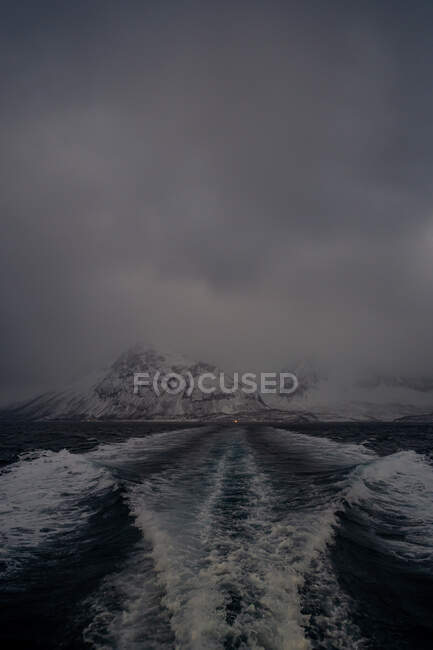 High angle of trail on foam waves left by water transport driving from solitary light on highland snowy shore under gray cloudy sky at twilight in Lofoten — Stock Photo