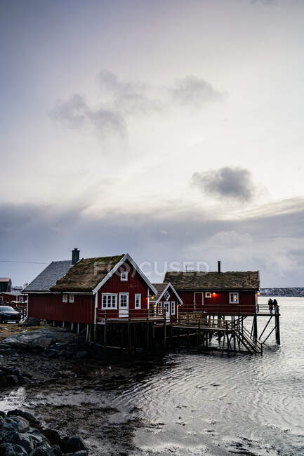 Pequeñas casas de campo rojas con muelles de madera en la playa rocosa del estrecho con agua tranquila en la noche fría en Noruega - foto de stock