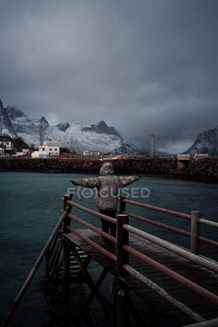 Back view of tourist in warm clothes and hood standing with arms outstretched on wooden pier against calm water of in fjord with township and snowy foggy mountains in Norway — Stock Photo