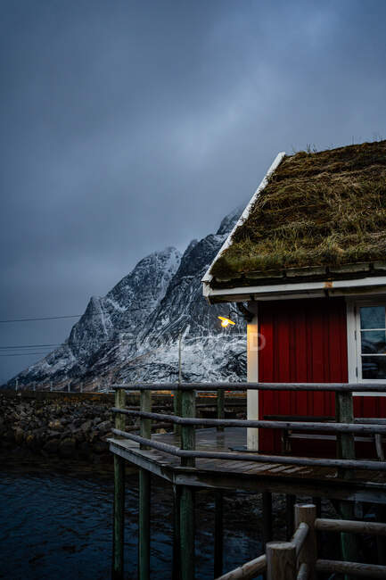 Cottage com paredes listradas vermelhas e molduras brancas da janela no cais de madeira na costa estreita contra o município nevado no foothill no dia nublado do inverno em Lofoten — Fotografia de Stock