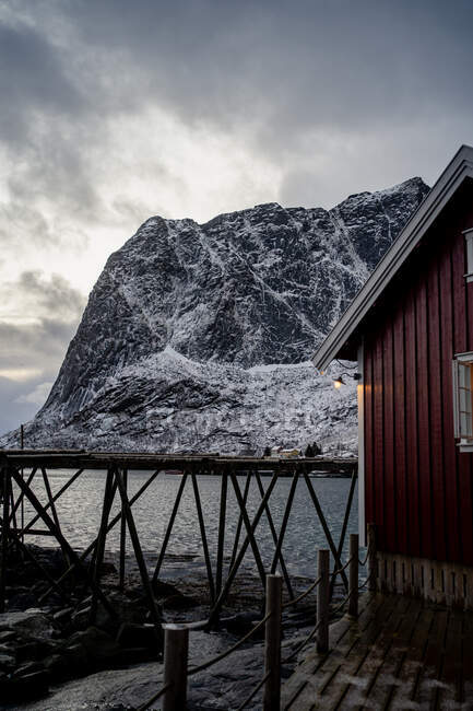 Cottage con pareti a strisce rosse e finestre bianche sul molo di legno sulla riva dello stretto contro la città innevata ai piedi in inverno giornata nuvolosa in Lofoten — Foto stock