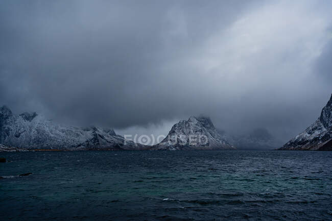 Vue à couper le souffle de l'eau de mer bleu ondulé contre les crêtes de montagne enneigées sur le rivage sous un ciel gris nuageux en hiver en Norvège — Photo de stock
