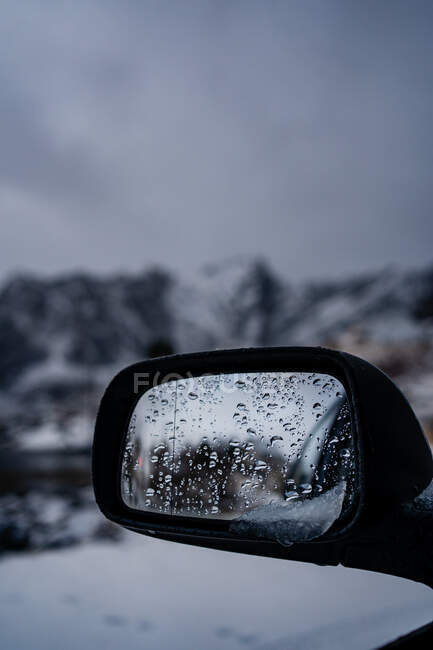 Мокрое боковое зеркало заднего вида современного черного авто с тающим снегом против размытой снежной возвышенности под серым облачным небом зимой в Норвегии — стоковое фото
