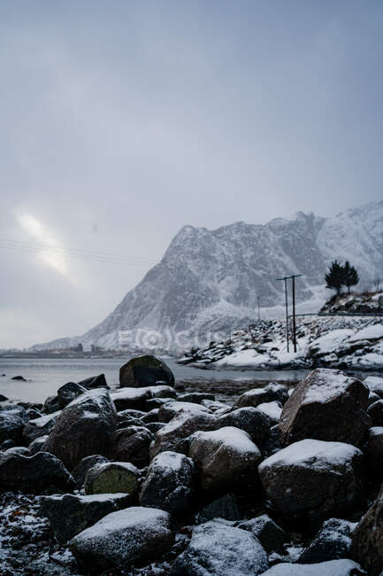 Paisagem pitoresca de praia com pedregulhos nevados contra a água do mar calma e crista montesa abaixo do céu nublado cinza em Lofoten — Fotografia de Stock