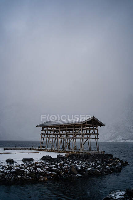 Construção solitária com pilares de madeira e telhado cinza na praia de pedra lavando por água conturbada contra cumes de montanha nebulosos em tempo nublado no porto na Noruega — Fotografia de Stock