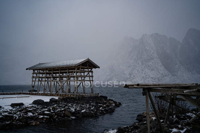 Одинокая конструкция с деревянными колоннами и серой крышей на каменном пляже, омываемая беспокойной водой против туманных горных хребтов в пасмурную погоду в гавани Норвегии — стоковое фото
