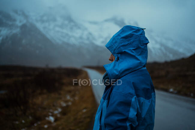 Back view of relaxed unrecognizable person in blue warm clothes and hoodie standing on asphalt road going to snowy foggy mountains in Lofoten — Stock Photo