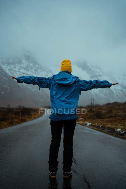 Vista posterior de la mujer sin rostro en ropa de abrigo azul y sombrero de gorro amarillo vivo disfrutando de idilio y frescura mientras está de pie con los brazos extendidos en el camino de asfalto que va a las cordilleras nevadas de niebla en Noruega - foto de stock