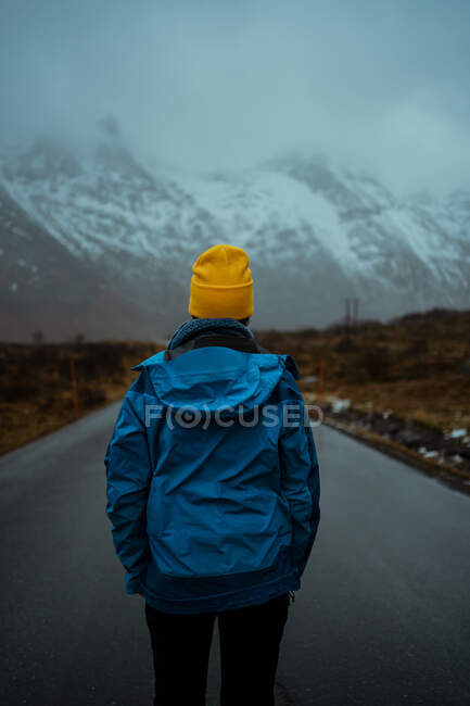 Повернення до розслабленої непізнаної людини в синьому теплому одязі і яскраво-жовтий капелюх біні, що стоїть на асфальтній дорозі йде снігом туманні гори в Лофотені. — стокове фото