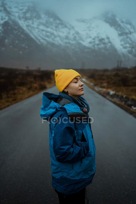 Vista laterale di felice donna rilassata in abiti caldi blu e cappello giallo brillante godendo la vita mentre in piedi con gli occhi chiusi su strada asfaltata andando a montagne nebbiose nevose in Lofoten — Foto stock