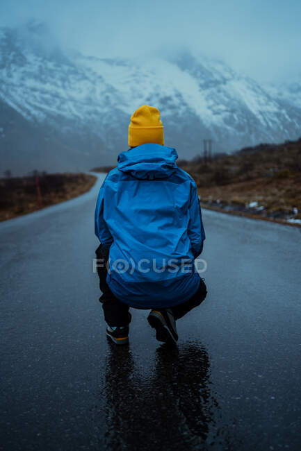 Rückansicht einer entspannten, unkenntlich gemachten Person in blauer warmer Kleidung und hellgelber Mütze, die auf einer Asphaltstraße kauert, die zu verschneiten nebligen Bergen auf den Lofoten führt — Stockfoto