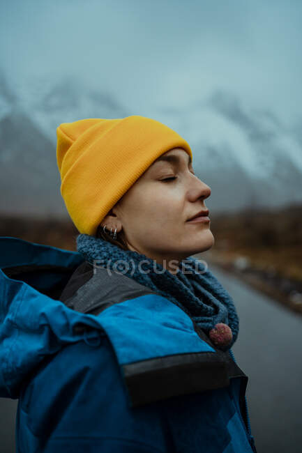 Side view of happy relaxed woman in blue warm clothes and bright yellow hat enjoying life while standing with eyes closed on asphalt road going to snowy foggy mountains in Lofoten — Stock Photo