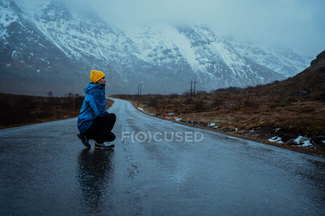 Vista laterale della donna rilassata in abiti caldi blu e cappello beanie giallo brillante godendo la vita mentre accovacciato guardando lontano sulla strada asfaltata andando a montagne nebbiose nevose in Lofoten — Foto stock